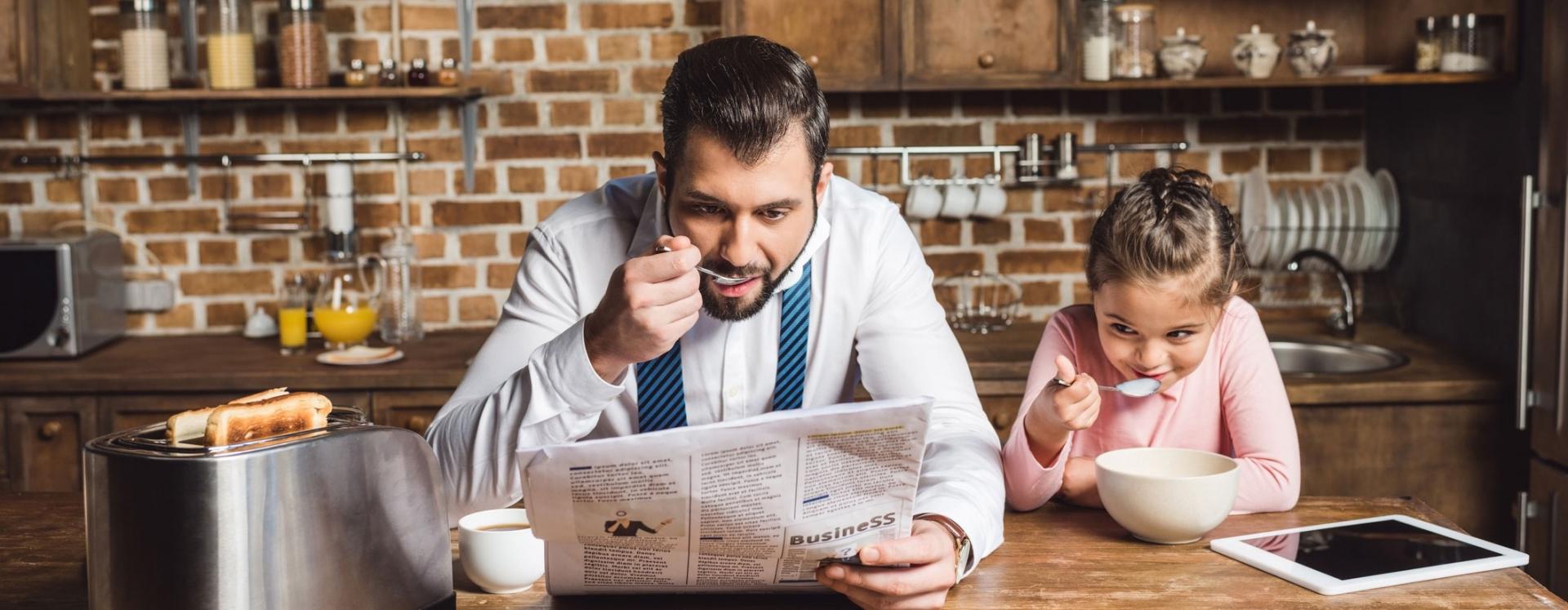 Father Reading Newspaper While Eating Bowl Of Cereal With Daughter