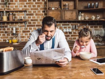 Father Reading Newspaper While Eating Bowl Of Cereal With Daughter