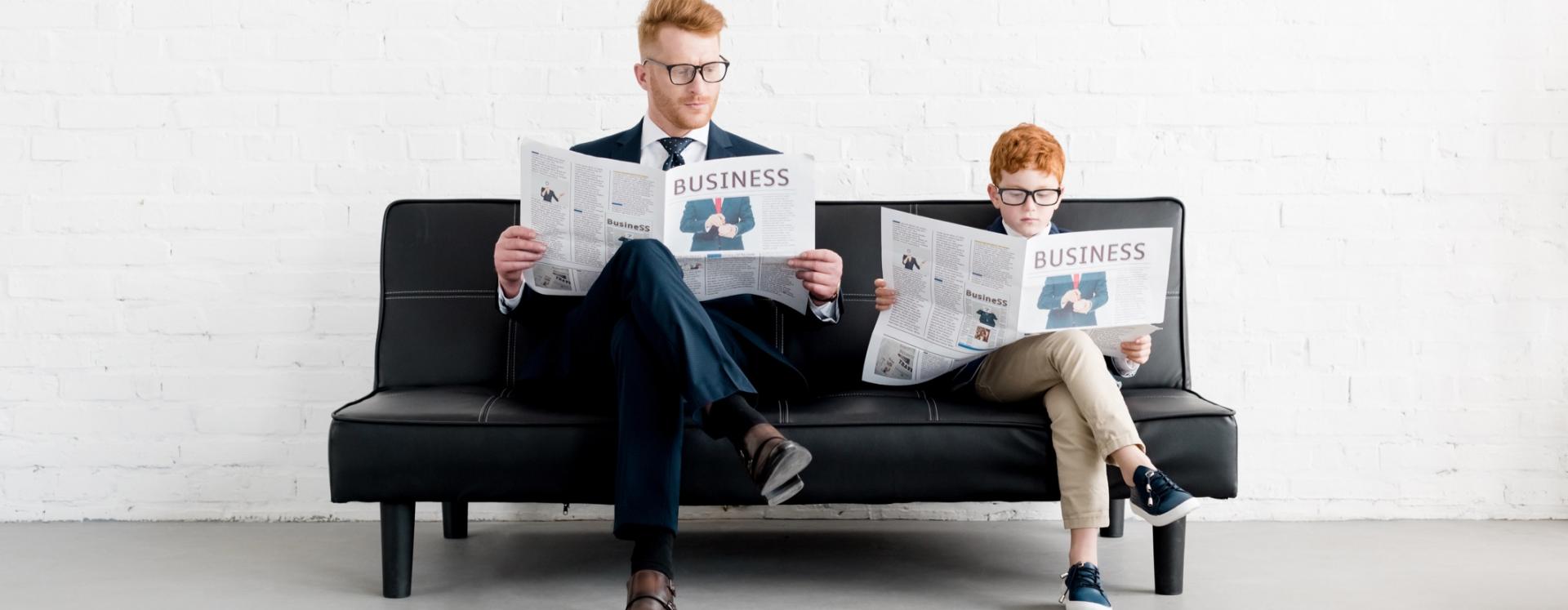 Man And Young Boy Sitting On Sofa Each Reading A Business Newspaper
