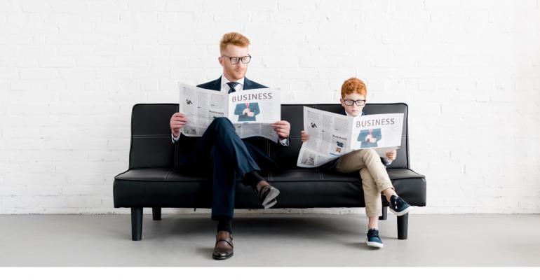 Man And Young Boy Sitting On Sofa Each Reading A Business Newspaper