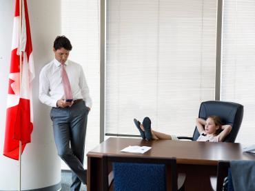 Justin Trudeau Standing Looking At Mobile Phone With Daughter Sitting At his Desk With Feet On Table
