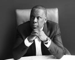 Black And White Photo Of Bruny Surin Sitting Down With Both Hands Under His Chin