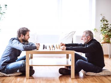 Young Man And Older Man Facing Each Other Sitting Down Playing Check