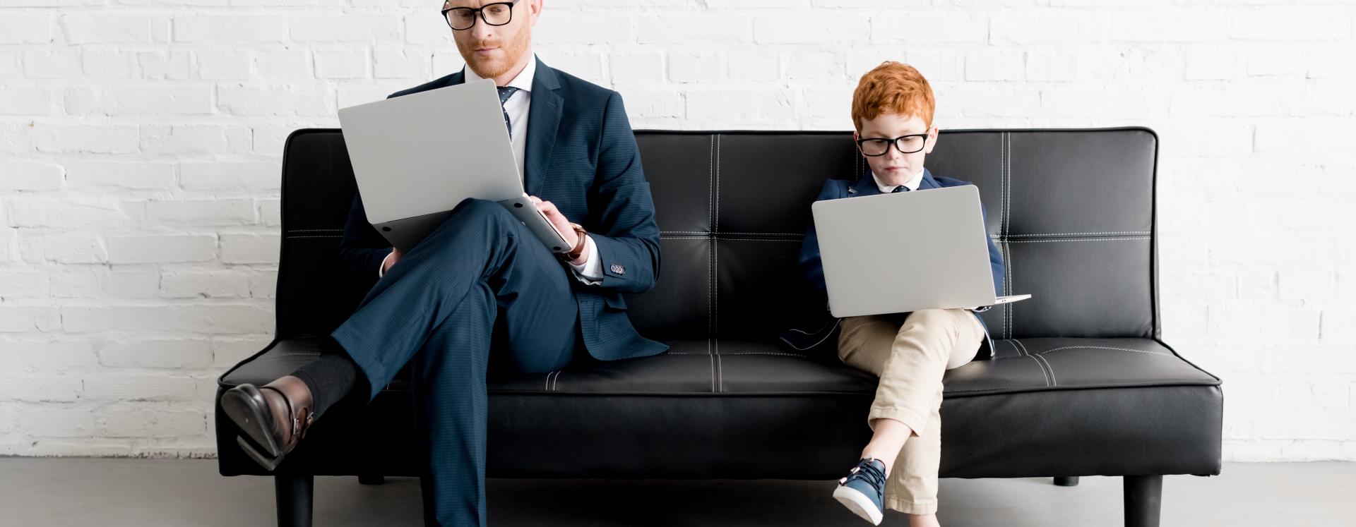 Business Man and Young Boy In Suit Sitting On Sofa With Their Laptops