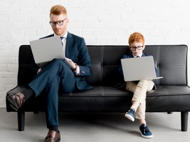 Business Man and Young Boy In Suit Sitting On Sofa With Their Laptops
