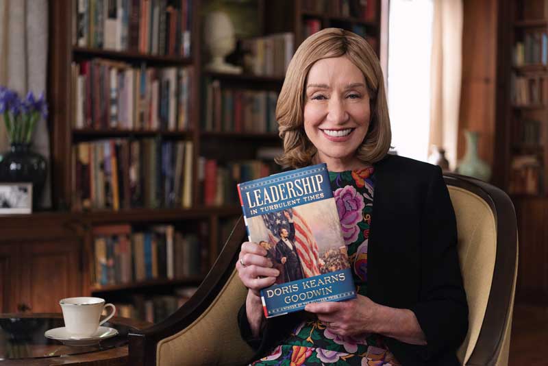 Doris Kearns Sitting On Chair Smiling And Holding Book Leadership In Turbulent Times